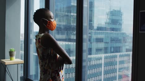 thoughtful african american woman wearing face mask looking out of window at modern office