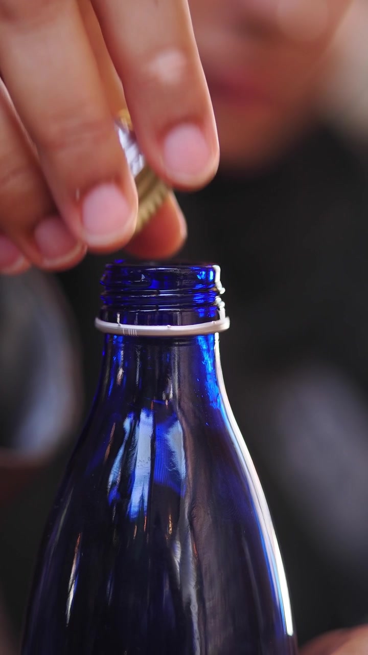Main dévissant un bouchon métallique d'une bouteille en verre bleu