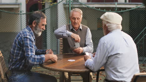 Hombre-Mayor-Jubilado-Jugando-A-Las-Cartas-Con-Sus-Dos-Mejores-Viejos-Amigos-Vecinos-En-El-Patio-Al-Aire-Libre