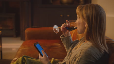 woman at home sitting on sofa with fire with blue screen mobile phone holding glass of wine