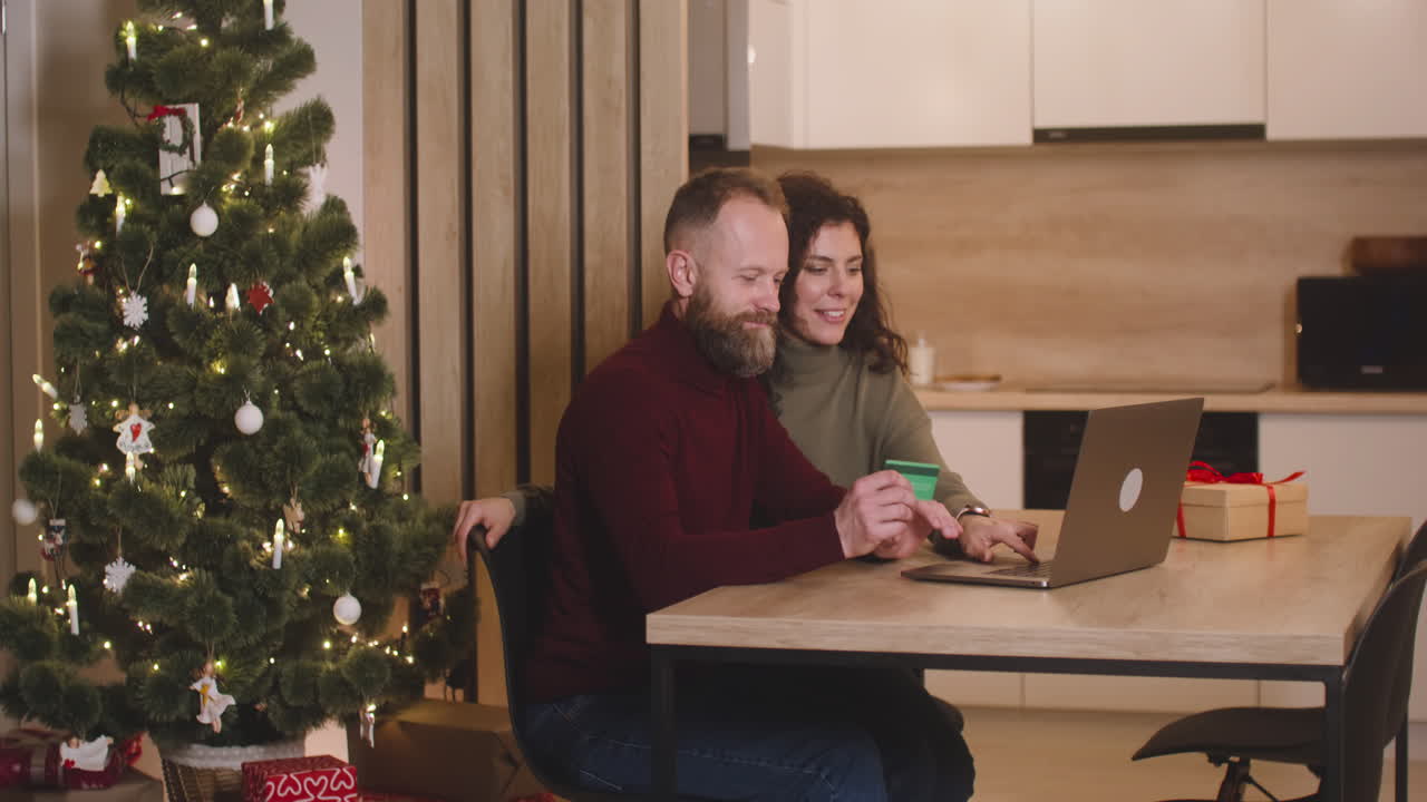 vista lateral de una pareja comprando en línea con una tarjeta de crédito usando una computadora portátil sentada en una mesa cerca de un regalo en una habitación decorada con un árbol de navidad