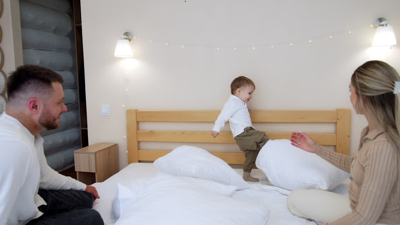 Happy Caucasian parents sit on the double bed looking at their adorable baby. Toddler kid kicks the pillows with the foot.