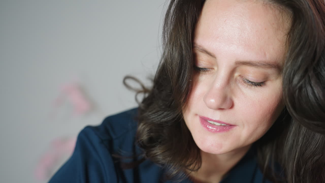 Female cosmetologist with bright face and focused expression working on client wearing navy uniform with long natural hair framing gentle smile in serene spa clinic with soft lighting equipment