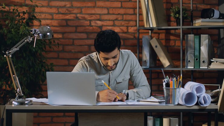 Young Arabian Architect Drawing Some Plans And Looking For Example On The Laptop Screen In His Modern Office Room