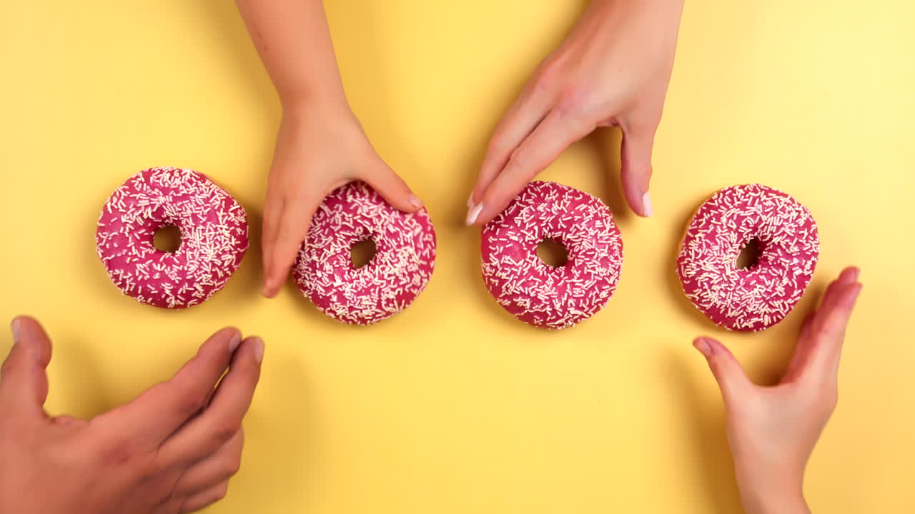 Topview of family taking four pink colorful donuts on light yellow background. Family grab delicious donut at same time. Pink glazed with sprinkles. Filmed in Prores, Topview, Slowmotion