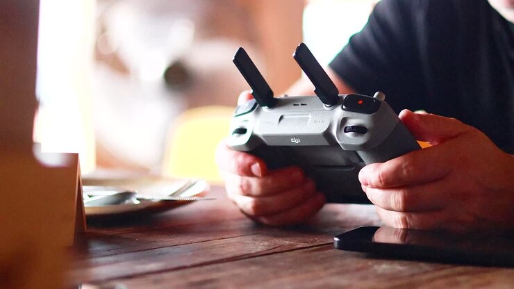Mani azionano un controller per drone su un tavolo di legno in un ambiente di caffè con luce soffusa, creando un'atmosfera concentrata e calma.