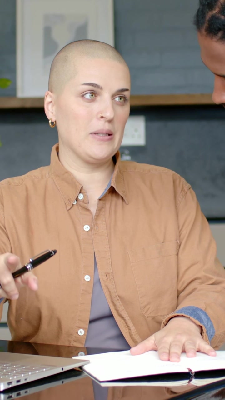 Video verticale: Donna con top marrone che scrive, stimolata da un suggerimento sul quaderno, mentre parla a una riunione, con laptop e penna.