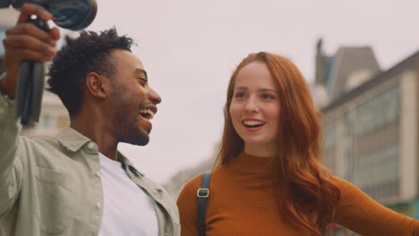 young couple travelling through city together vlogging to video camera on handheld tripod