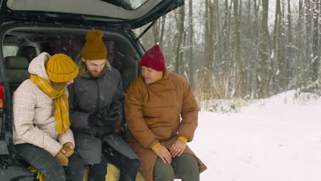 bärtiger mann sitzt mit zwei freunden im kofferraum und schaut an einem wintertag auf das handy