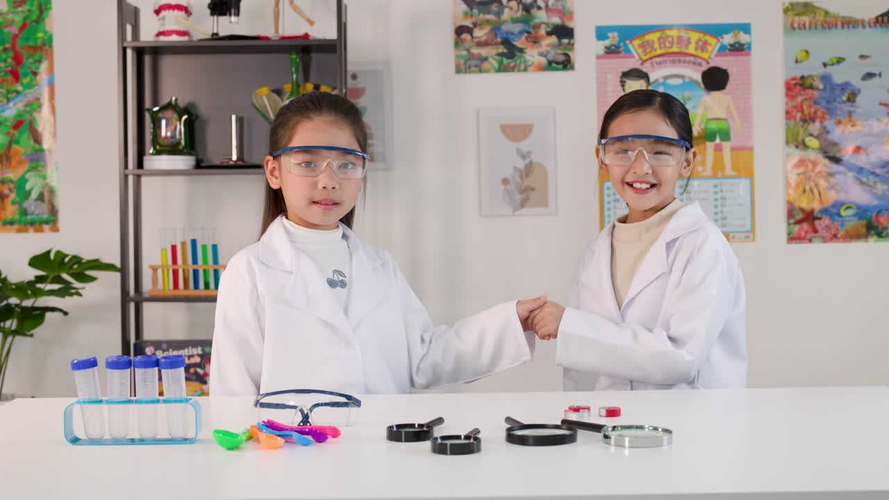 Dua gadis muda Asia mengenakan jas lab dan kacamata keselamatan berjabat tangan di ruang kelas