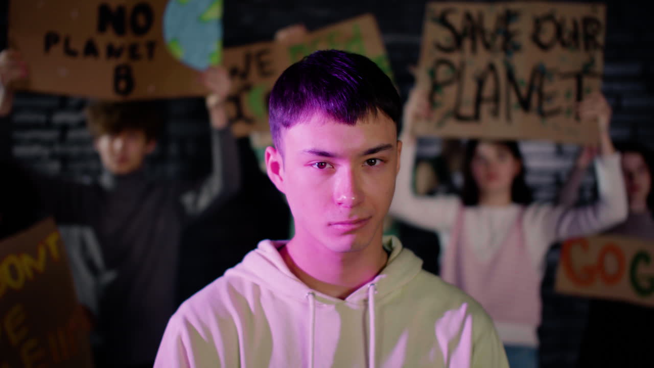 그의 친구들이 플래카드를 들고 그 뒤에서 항의하는 동안 카메라를 바라보는 진지한 젊은 남성 환경 운동가의 초상화