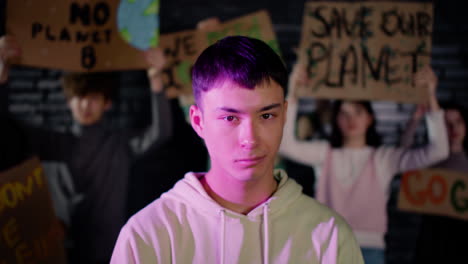 retrato de un joven activista ambiental masculino serio mirando la cámara mientras sus amigos sostienen pancartas y protestan detrás de él