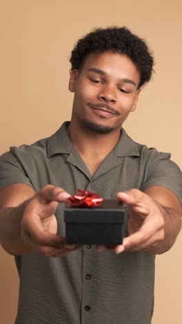 happy man giving gift box and looking at camera in beige studio