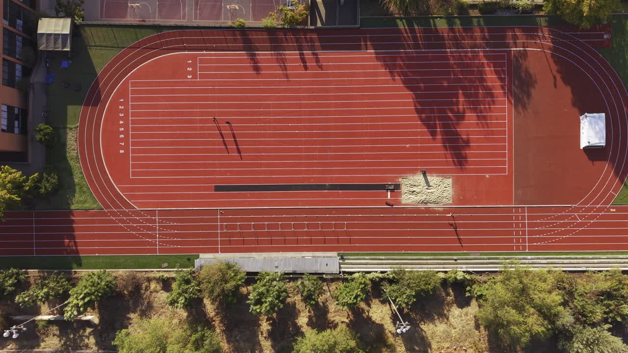 Fugleperspektiv af en person, der løber på en lys rød løbebane ved Colegio Cumbres, med en længdespringsgrav og omkringliggende træer. Chile, 4K.