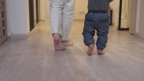 rear view of a mother helping her baby to take his first steps in the living room at home