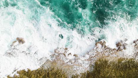 Aerial-descend-as-green-rocky-rough-ocean-waves-crash-on-jagged-drop-off-into-sea-spreading-whitewash