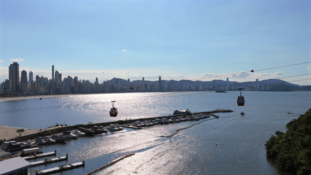 Parque Unipraias cable car over Molhe da Barra Sul with marina and boats in Barra Sul, Balneário Camboriú, Santa Catarina, Brazil, with Praia Central skyline and Serra do Mar in background drone orbit