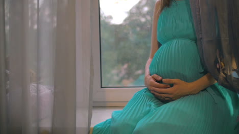 pregnant woman sitting on windowsill and embracing belly