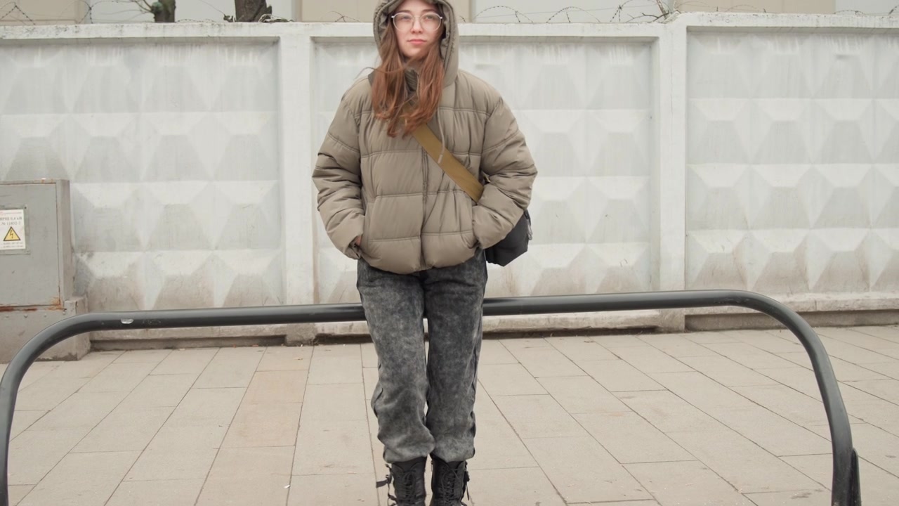 Giovane donna che cammina lungo il marciapiede, si ferma per guardare la fotocamera con un piumino con cappuccio, mani nelle tasche, stivali robusti, sfondo di muro neutro e rastrelliera per biciclette in metallo in primo piano, umore fiducioso, stile street