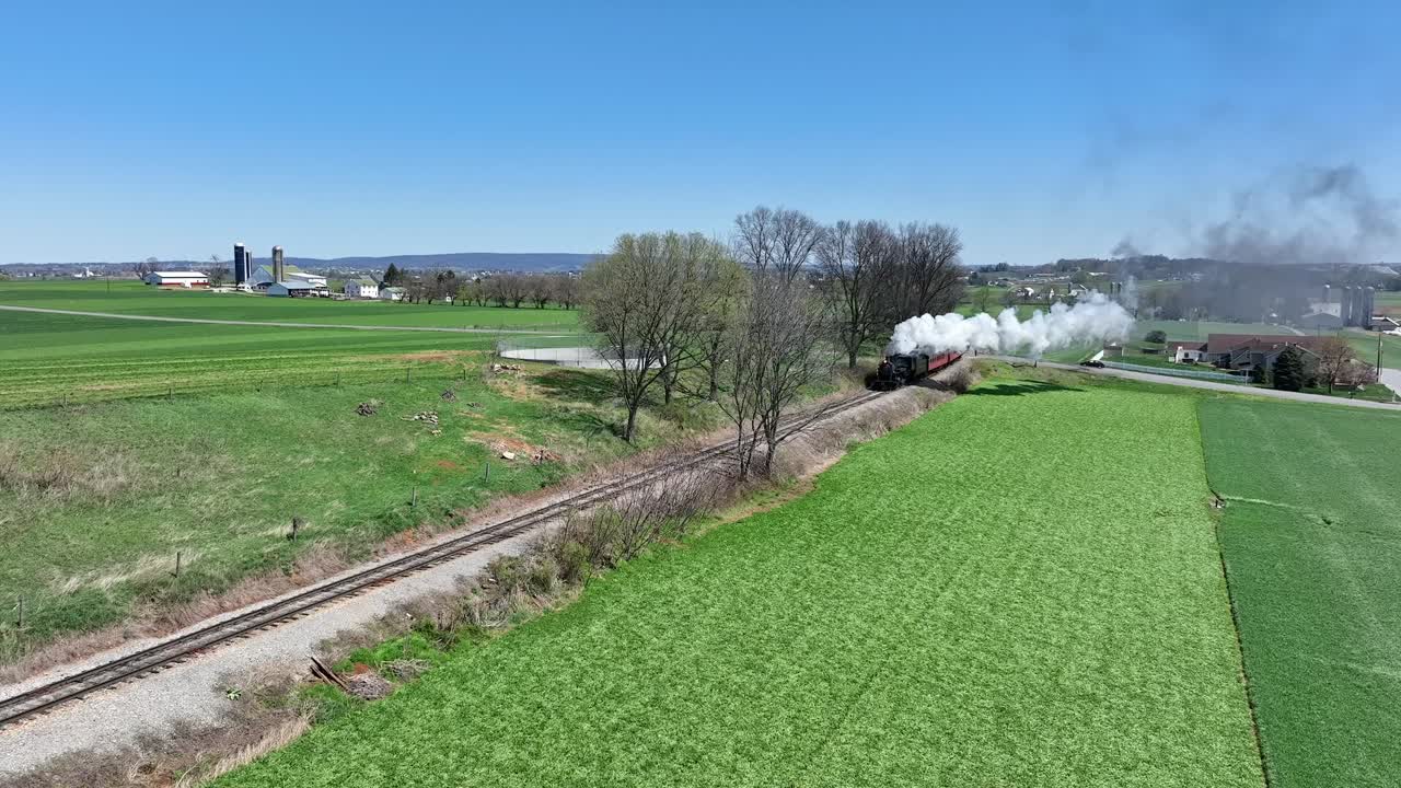 A steam train moves along tracks lined by lush green fields under a clear blue sky. The scene captures the beauty of nature and the charm of countryside landscapes.