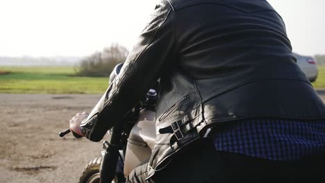 Vista-Lateral-De-Un-Hombre-Con-Casco-Negro-Y-Chaqueta-De-Cuero-Montando-Motocicleta-En-Una-Carretera-Asfaltada-En-El-Campo.-Día-Soleado