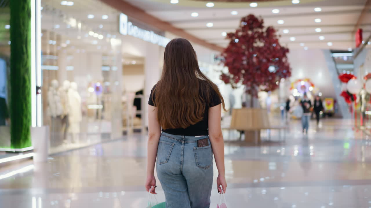 청바지와 검은색 을 입은 여자가 활기차고 잘 조명 된 쇼핑몰에서 화려한 쇼핑 가방을 들고 다른 쇼핑객과 반이는 타일과 함께 뒷면