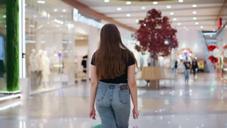 back view of a lady in jeans and black top carrying colorful shopping bags in a bustling, well-lit mall with other shoppers and reflections on glossy tiles