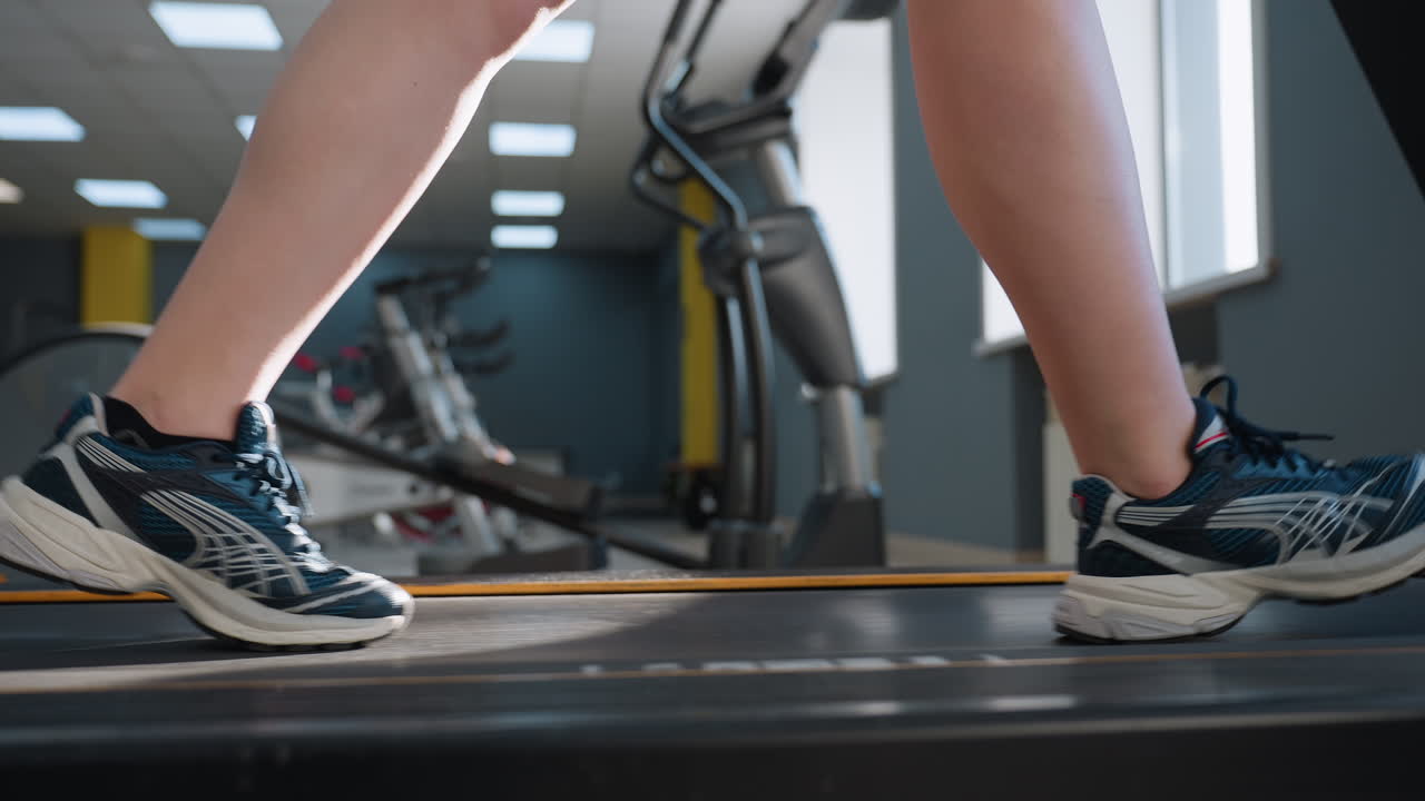 side leg view of woman wearing canvas sneakers walking on treadmill belt in bright gym studio under overhead lighting with exercise bikes and elliptical trainer visible through window