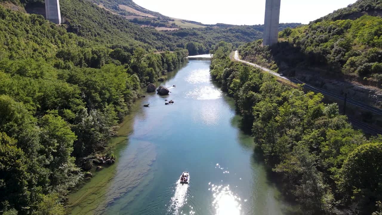Aerial shot following a boat up the green river with car passing by on the road and tall viaduct in the back. Small boat moving up the river with gorgeous reveal of viaduct and beautiful river valley
