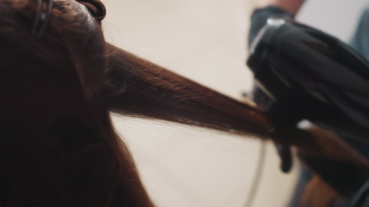 Nærbillede af føntørring med handsker. Professionel stylist sektionerer brunt hår og bruger en højvarme-tørrer til at glatte håret, hvilket skaber en glansfuld finish og kontrolleret bevægelse i et varmt studielys.