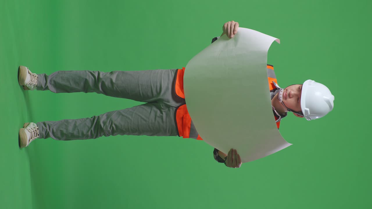 Full Body Of Asian Male Engineer With Safety Helmet Looking At Blueprint In His Hands And Looking Around While Standing In The Green Screen Background Studio