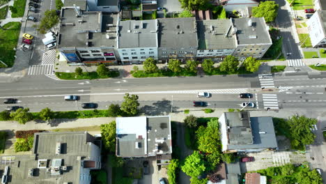 aerial top-down view of a busy city street, with buildings on both sides, cars moving along the road, and residential homes with green gardens