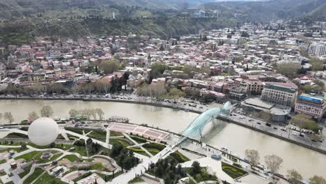 Aerial-shot-of-peace-bridge-Georgia-Tbilisi-city-center-river-cars-people-old-buildings