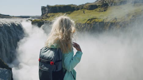 islandia. primer plano trasero de una mujer de cabello rubio de pie en el borde de un acantilado con una hermosa cascada de detifoss en el fondo