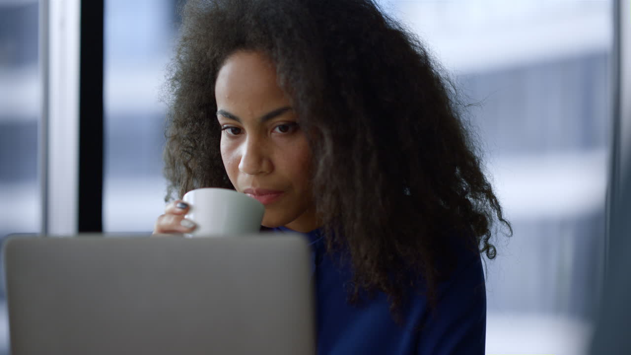 Serious african american woman executive working on laptop computer drinking morning coffee in home office. Portrait of focused business person reading digital device in workplace. Corporate concept.
