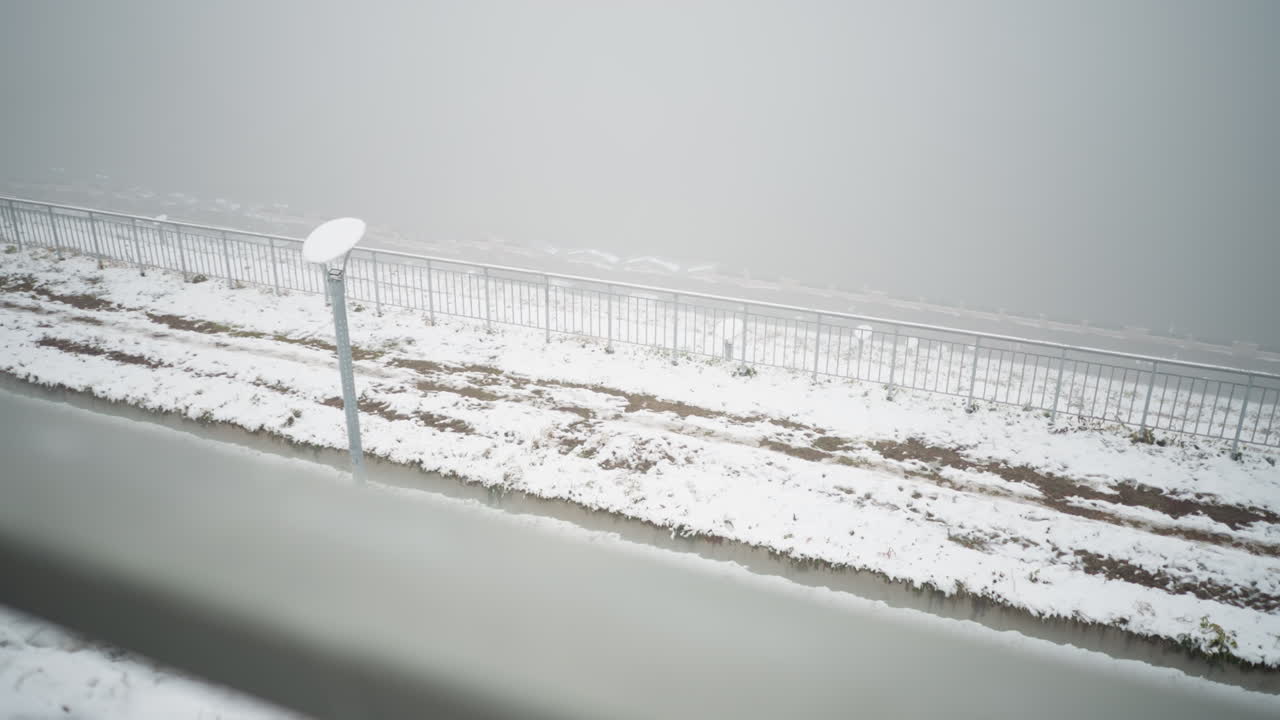 눈 인 풍경, 안개가 가득한 분위기, 금속 레일링, 조명 기둥, 그리고 희미하게 보이는 멀리 있는 특징, 땅은 눈 어리와 갈색 토양을 특징으로 하며, 평온하고 겨울의 장면을 만니다.