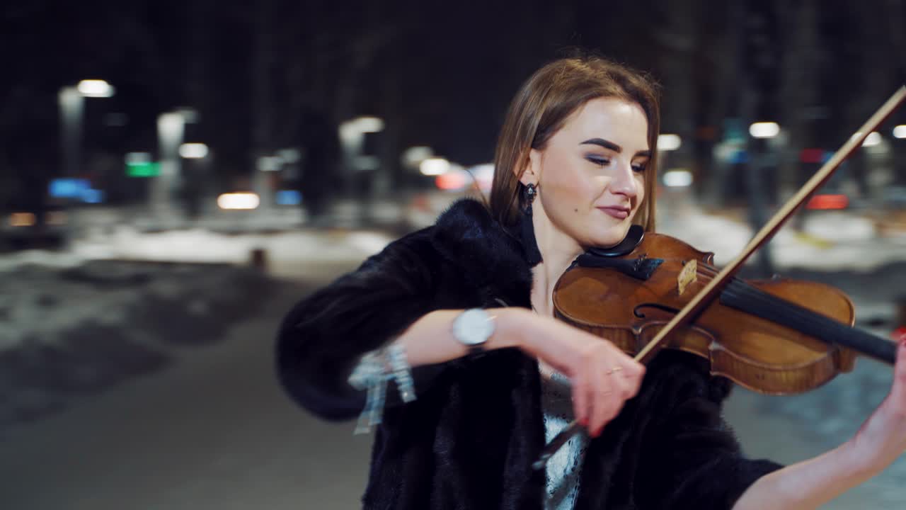 A pretty girl stands in the middle of the square and holds the violin with one hand, the bow with the other hand and performs the composition in the evening. Close-up.
