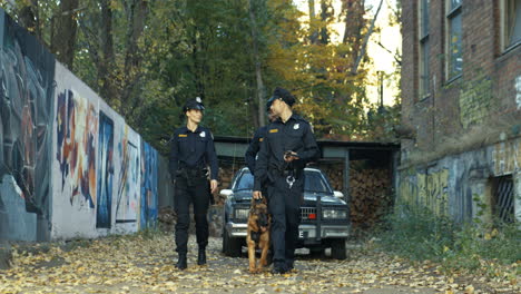 Three-Multiethnic-Police-Workers-Walking-The-Old-Lane-With-A-Shepherd-Dog-And-Searching-Something