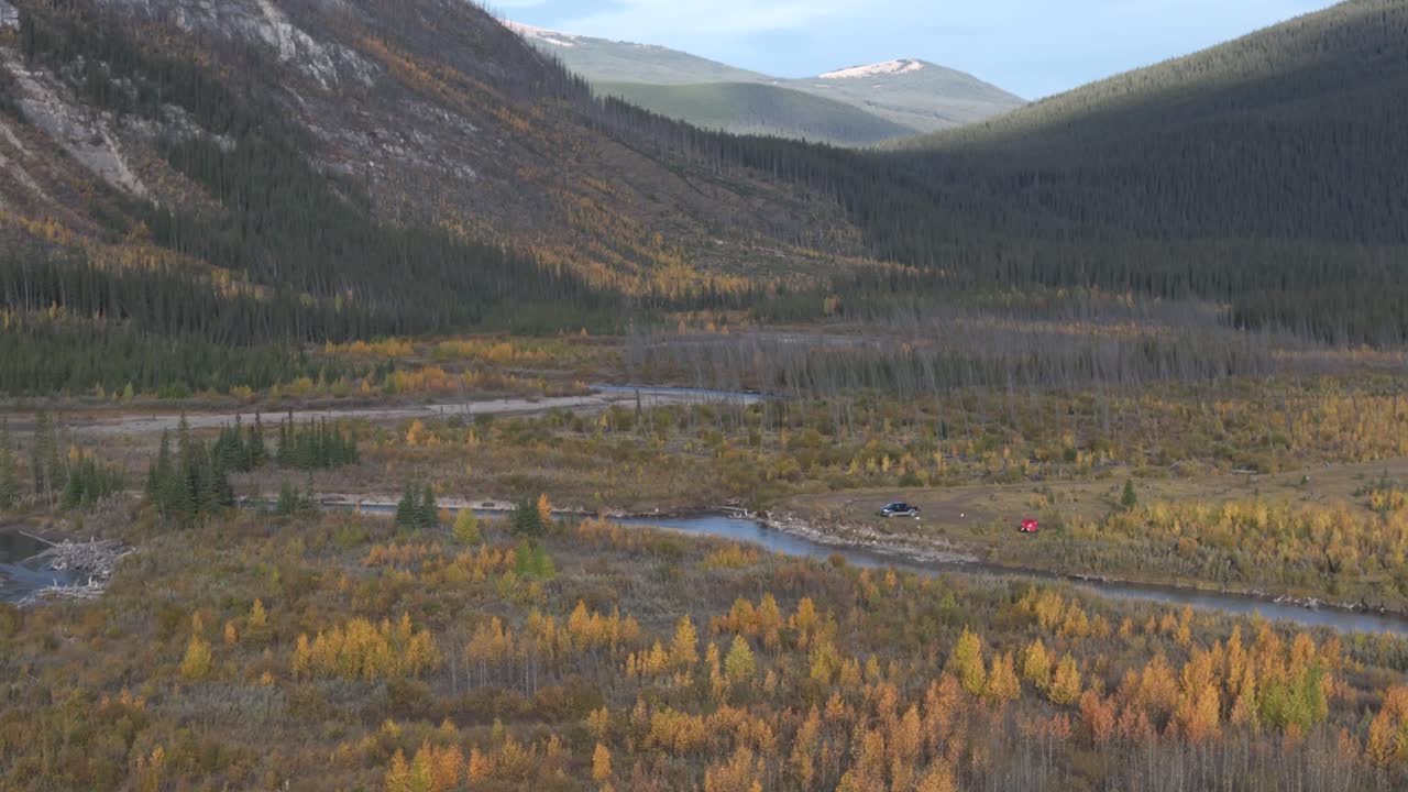 un campamento aislado con una tienda roja y un camión está retratado en un valle remoto a lo largo de un río serpenteante de las montañas rocosas
