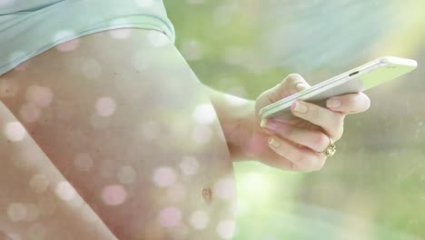 manchas de luz bokeh sobre la sección media de una mujer caucásica embarazada que usa un teléfono inteligente en el sol