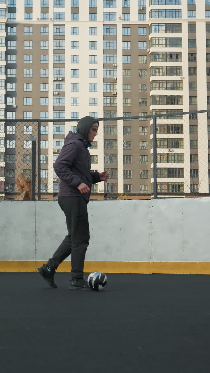 Man practicing soccer skills, controlling ball with precision while walking on outdoor sports ground, urban backdrop features high-rise buildings and goalpost