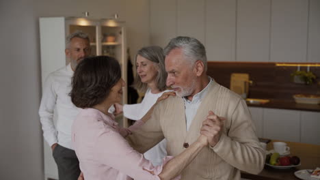 Two-Happy-Senior-Couples-Dancing-In-The-Kitchen,-While-On-Blurred-Background-A-Pretty-Elderly-Woman-Filming-Them-On-Mobile-Phone