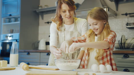 una chica adolescente linda rompiendo un huevo en el cuenco de vidrio con una hija adentro y su madre mirándola haciendo esto y controlando la cocina. retrato.