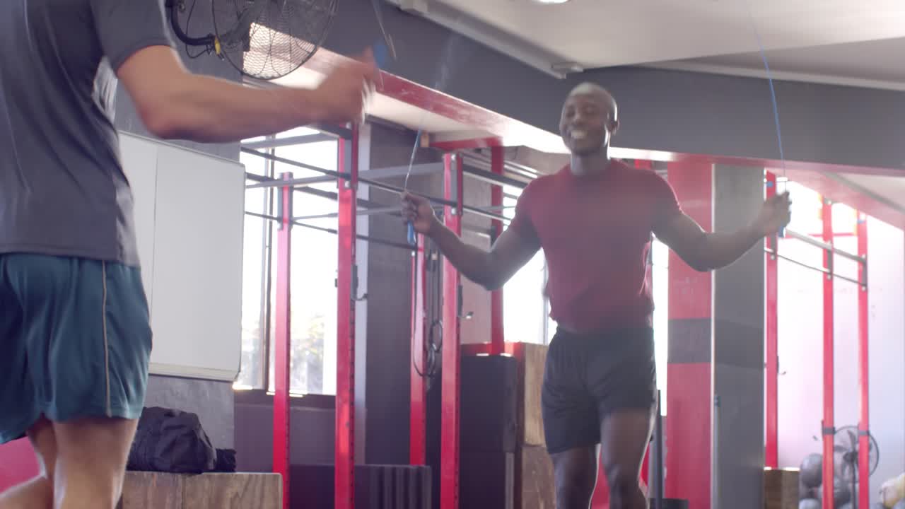 Diverse male duo holding jump rope overhead and synchronizing jumps in gym for cardio workout