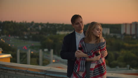 man and young pretty woman sitting on couch holding wine glasses and kissing on rooftop terrace at sunset