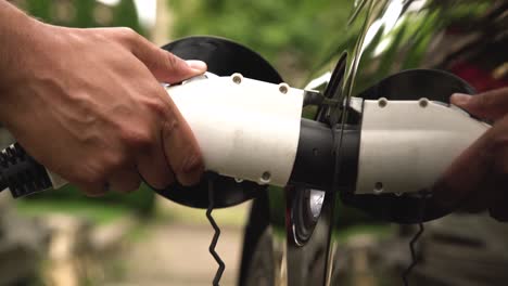 mans hand inserting charger plug into electric car in green environment background. new energy vehicle, nev is being loaded with electricity power. ecology, modern day cars