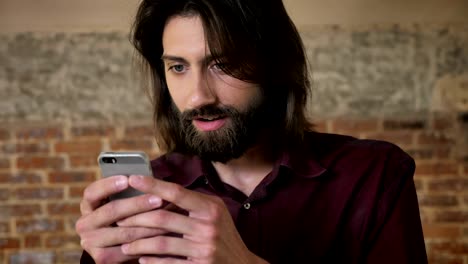 joven chocado hombre moreno con barba está viendo fotos en el teléfono inteligente, fondo de ladrillo, concepto de comunicación