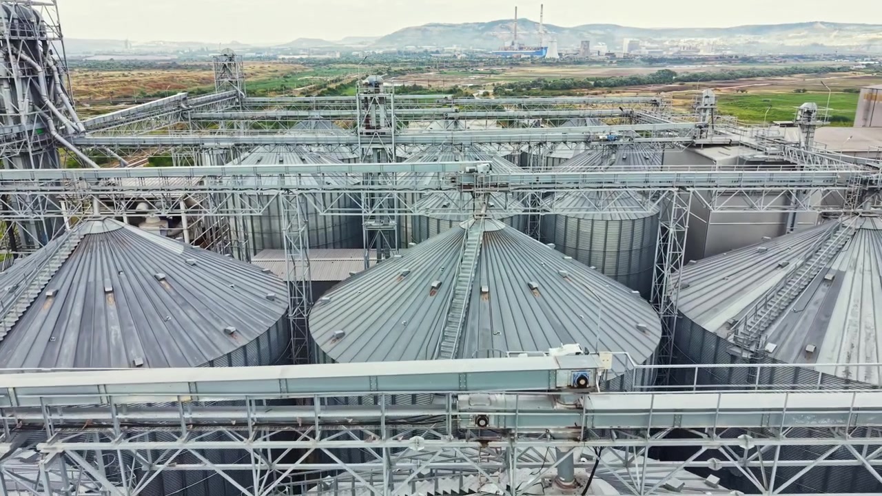 Industrial storage silos are seen from above in a farming area during daytime with mountains in the background