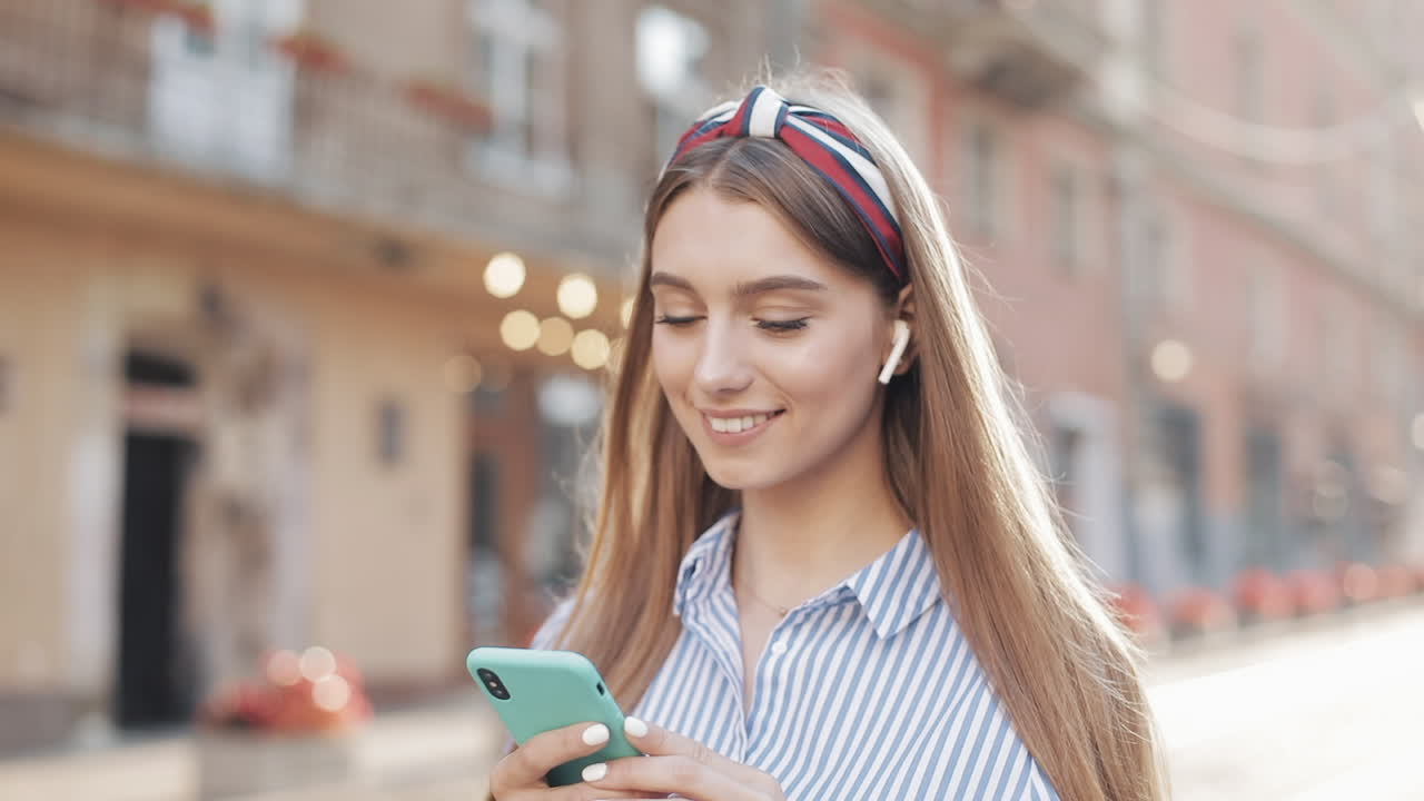 Giovane donna felice che usa uno smartphone e auricolari wireless in una strada cittadina.