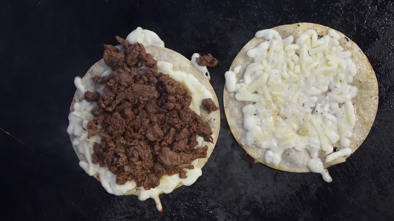 Nærbillede af to frisk tilberedte tortillas på en grill, den ene med asada og den anden med smeltet ost. Varm damp og enkel komposition skaber en ren baggrund af gadekøkken.